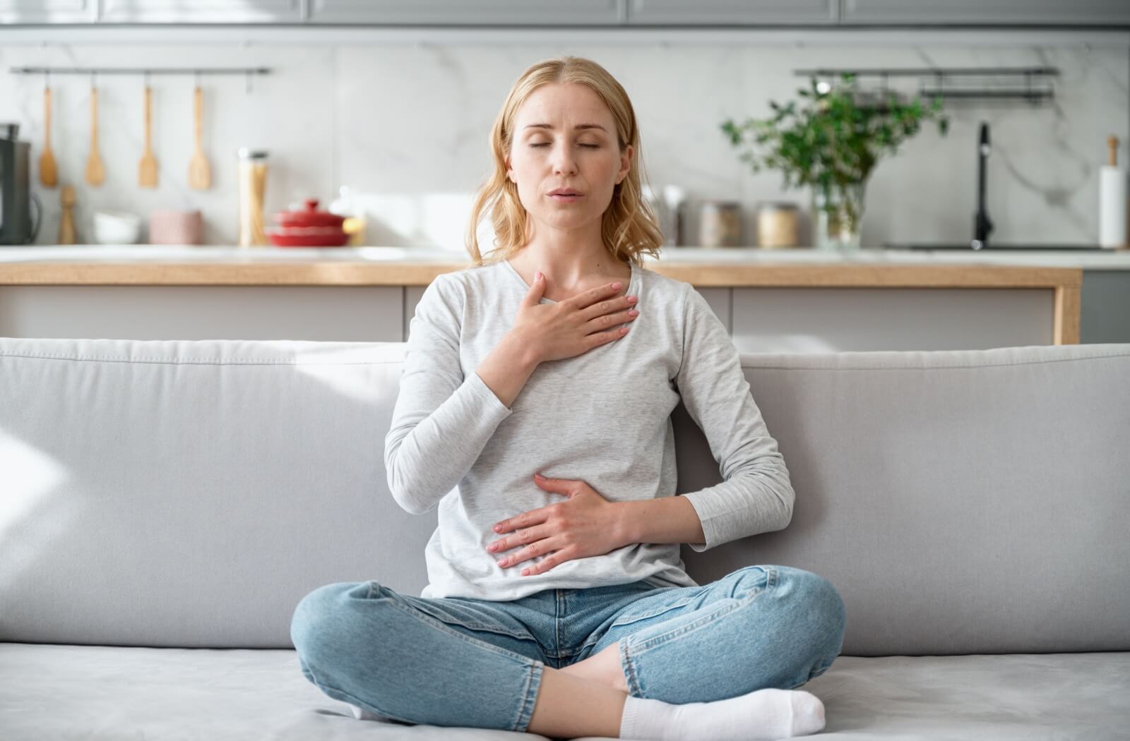 A person with eyes closed breathing while sitting on the couch with crossed legs and hands on their chest and stomach.