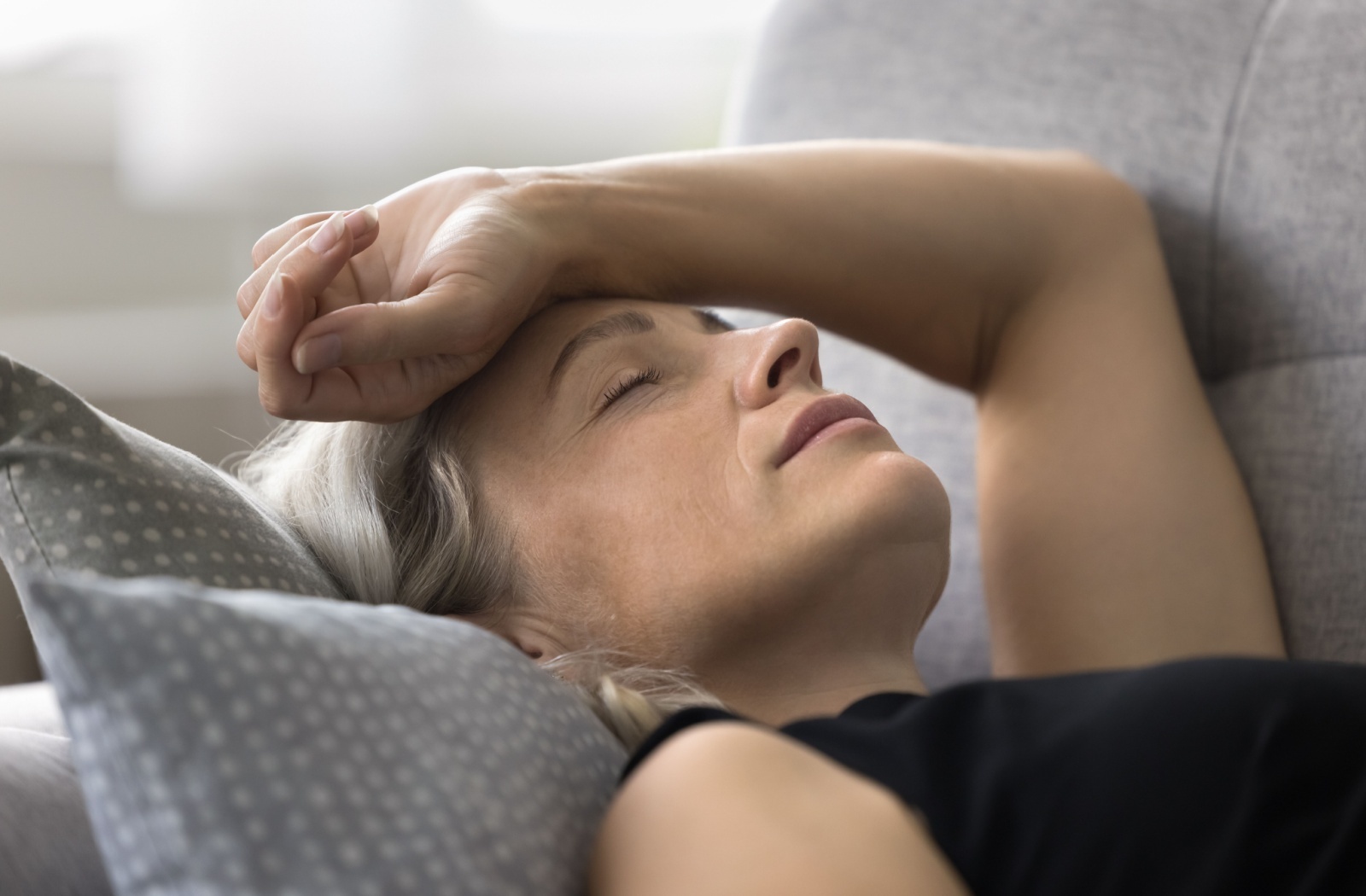 Person lying on couch with eyes closed and hand on forehead appearing in pain or distress