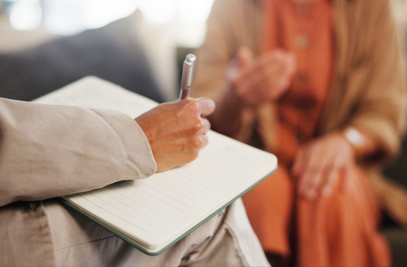 A hand of a psychologist writing in a notebook with a patient sitting across from them.